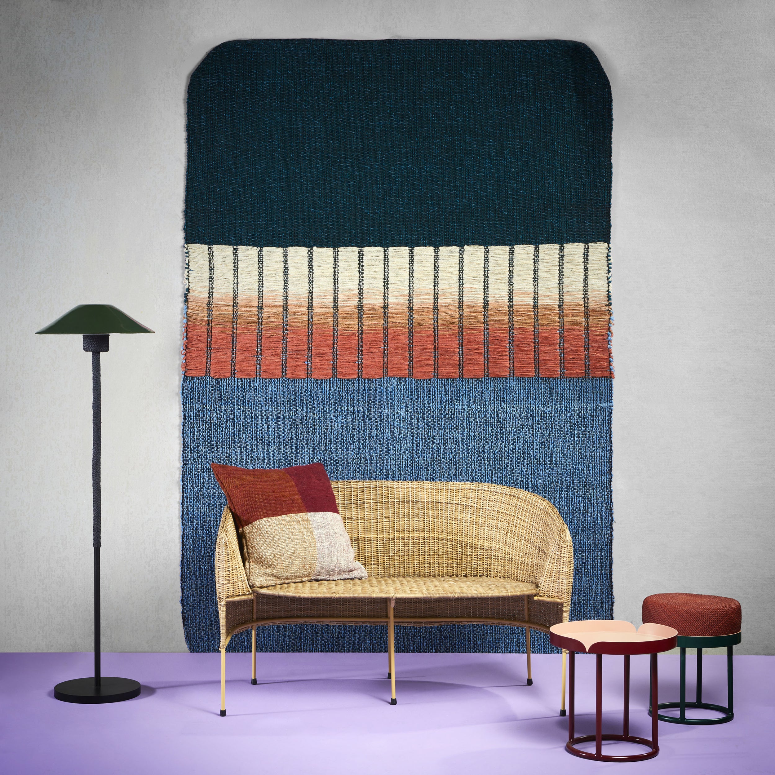 A stylish room boasts an Ames Balcones Rug on the light purple floor, a hand-woven tapestry with dark blue, beige, and rust stripes on the wall, a wicker bench with a multicolored cushion, a floor lamp, and three nesting tables with colorful tops.