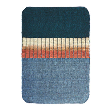 The Ames Balcones Rug is a hand-woven textile art piece with an elegant gradient: dark teal at the top, beige and orange stripes in the center, and light blue at the bottom. The edges are beautifully rounded.