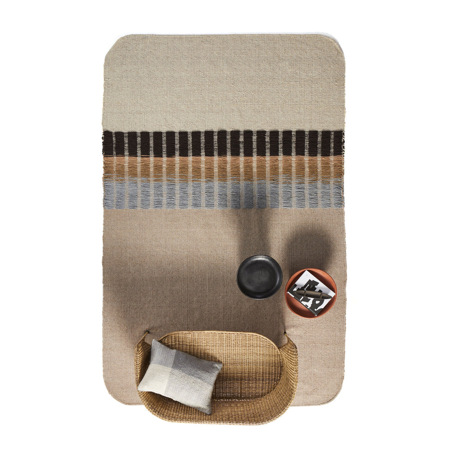 A top view of a bedroom setup highlights Ames Balcones Rug with geometric patterns. A wicker chair with a cushion and a small side table holding a book and black cup complete the cozy, modern decor.