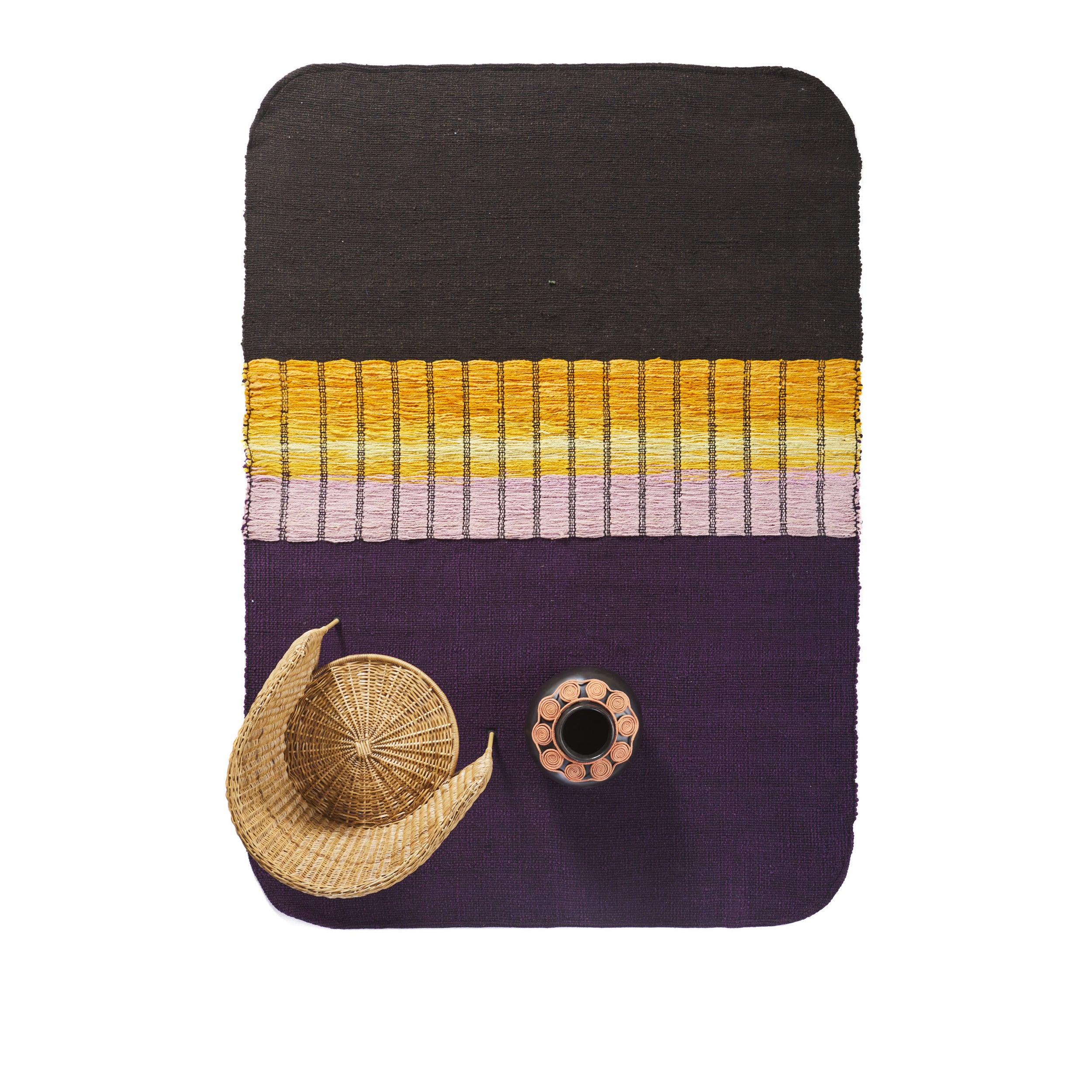 Amess Balcones Rug, a vibrant rectangular design in black, yellow, pink, and purple hues, is paired with a woven basket and circular tray for holding small items like a round cup in the center, inspired by Colombian cityscapes.