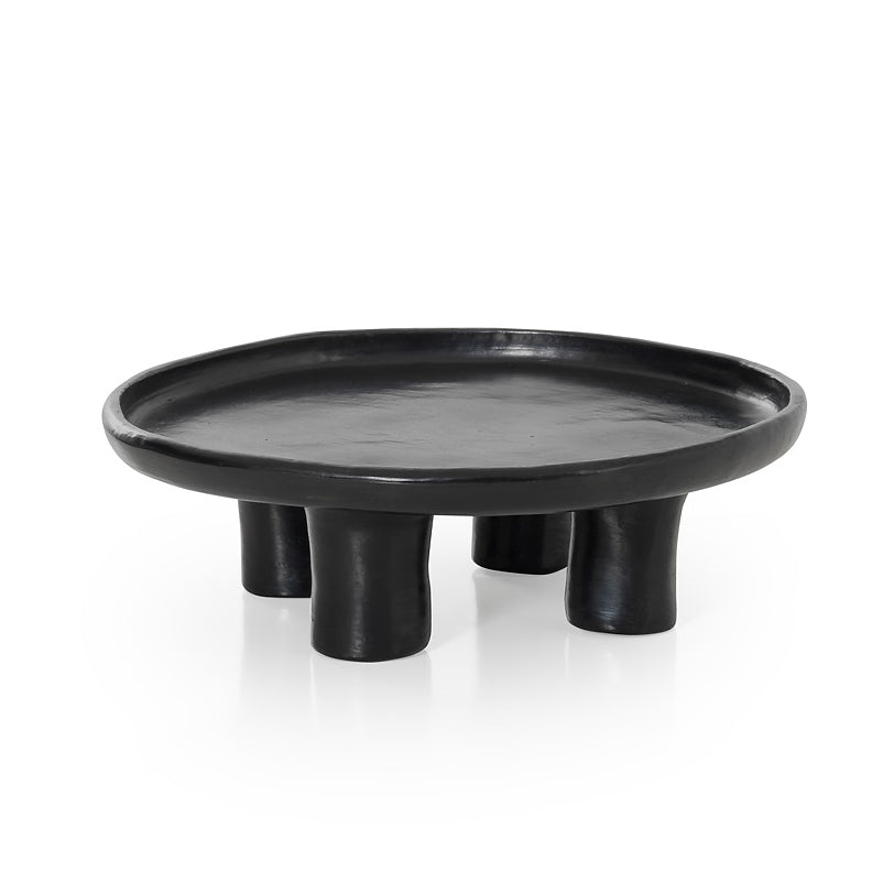 The Ames Barro Tray 1 by Ames, a round black modern coffee table with a raised edge and four thick cylindrical legs, displays handmade Colombian ceramics against a white background.