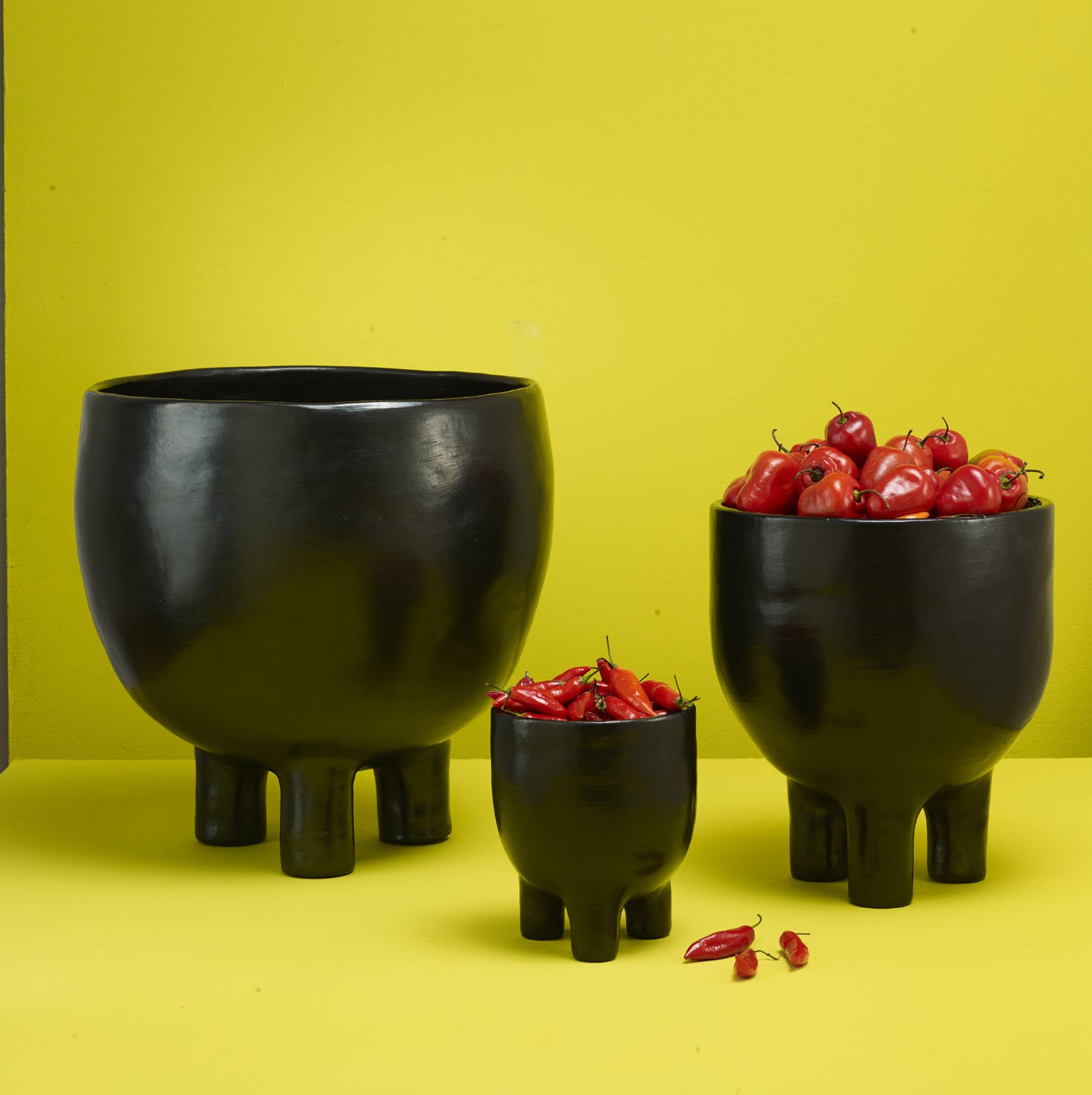 The Barro Pot 2 by Ames, consisting of three black ceramic bowls with short legs, stands against a bright yellow background. The smallest bowl contains red chilies, the medium one holds small red tomatoes, and the largest is empty.