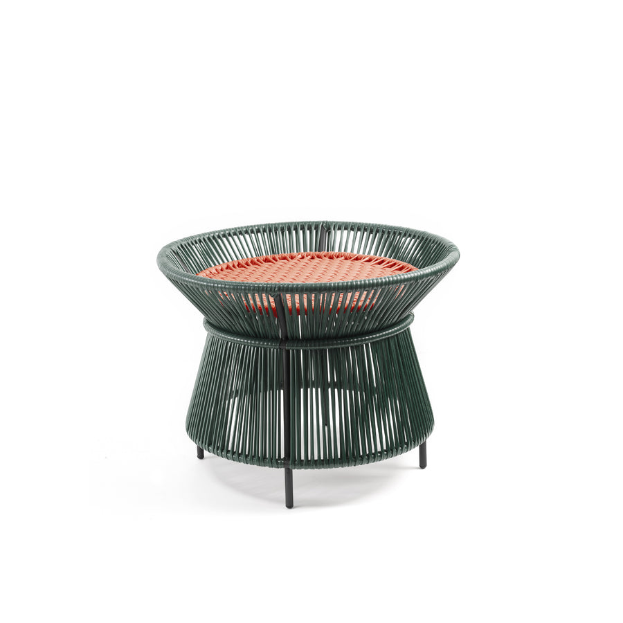 The Caribe Chic Basket Table by Ames is a small, round stool with a green wireframe structure and a red woven top made from recycled plastic cord. Its base slightly flares, standing on short black legs, combining modern minimalism with traditional Momposino weaving.