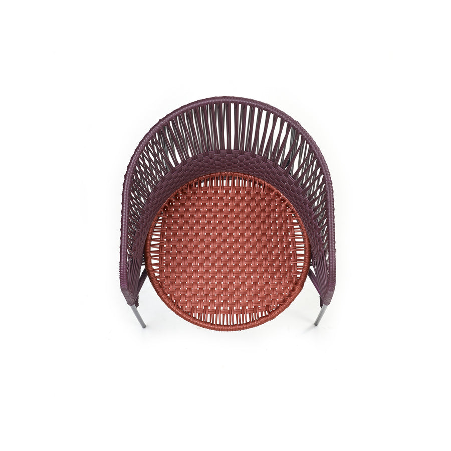 Top view of the Ames Caribe Chic Dining Chair by Sebastian Herkner, showcasing a complex woven pattern. The reddish-brown seat made from recycled plastic cord contrasts with a darker backrest, casting a subtle shadow on a white background.