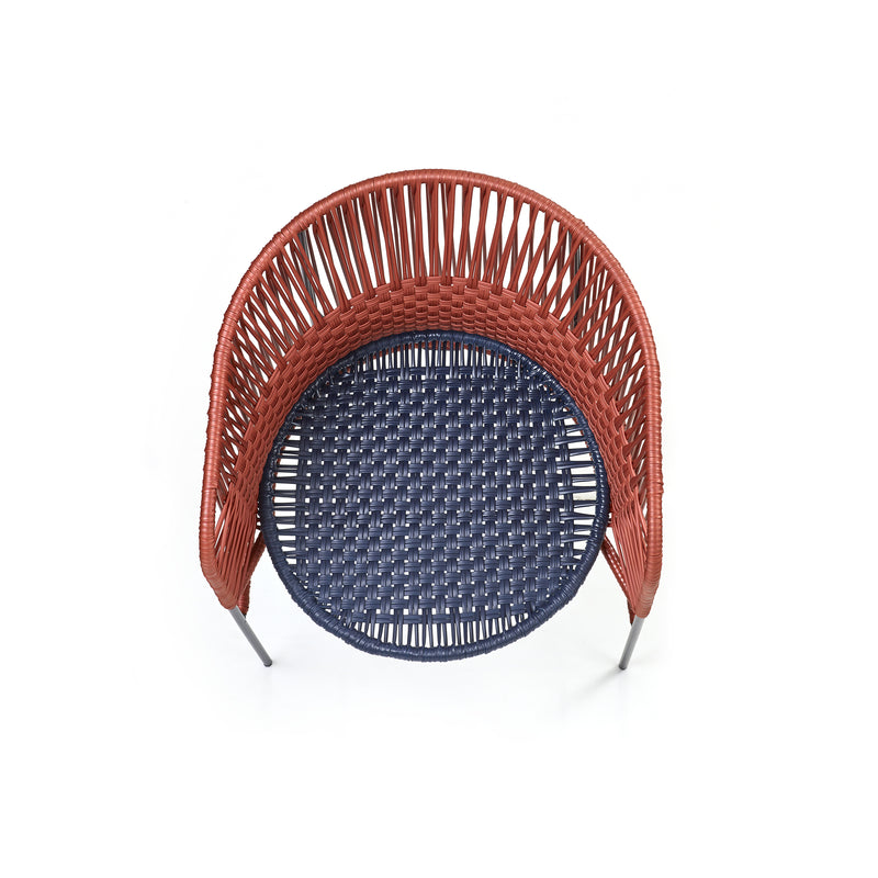 The Caribe Chic Dining Chair by Ames features a top-view wicker design with a round seat. It has reddish-brown woven backrest and sides, a dark blue seat made of recycled plastic cord, and thin metallic legs, all under the artistic direction of Sebastian Herkner.