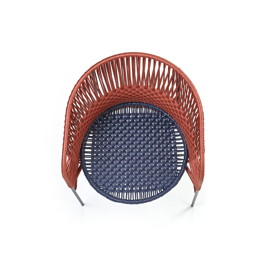 The Caribe Chic Dining Chair by Ames features a top-view wicker design with a round seat. It has reddish-brown woven backrest and sides, a dark blue seat made of recycled plastic cord, and thin metallic legs, all under the artistic direction of Sebastian Herkner.