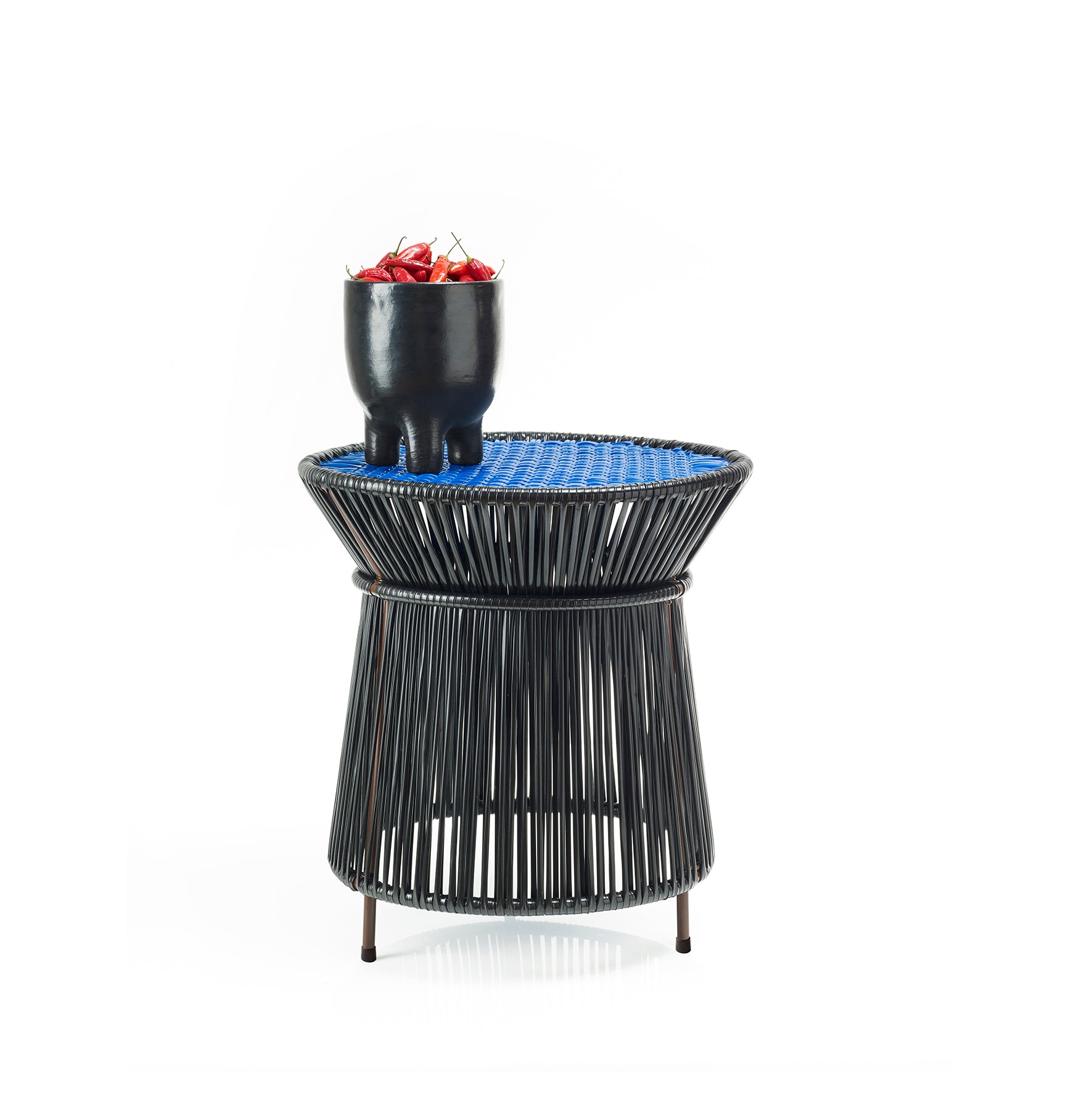 The Ames Caribe High Table by Ames, designed by Sebastian Herkner, features a blue woven top and black frame, displaying a small black pot with red berries—an elegant outdoor accent set against a plain white background.