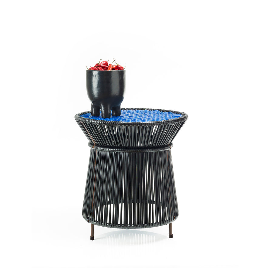 The Ames Caribe High Table by Ames, designed by Sebastian Herkner, features a blue woven top and black frame, displaying a small black pot with red berries—an elegant outdoor accent set against a plain white background.