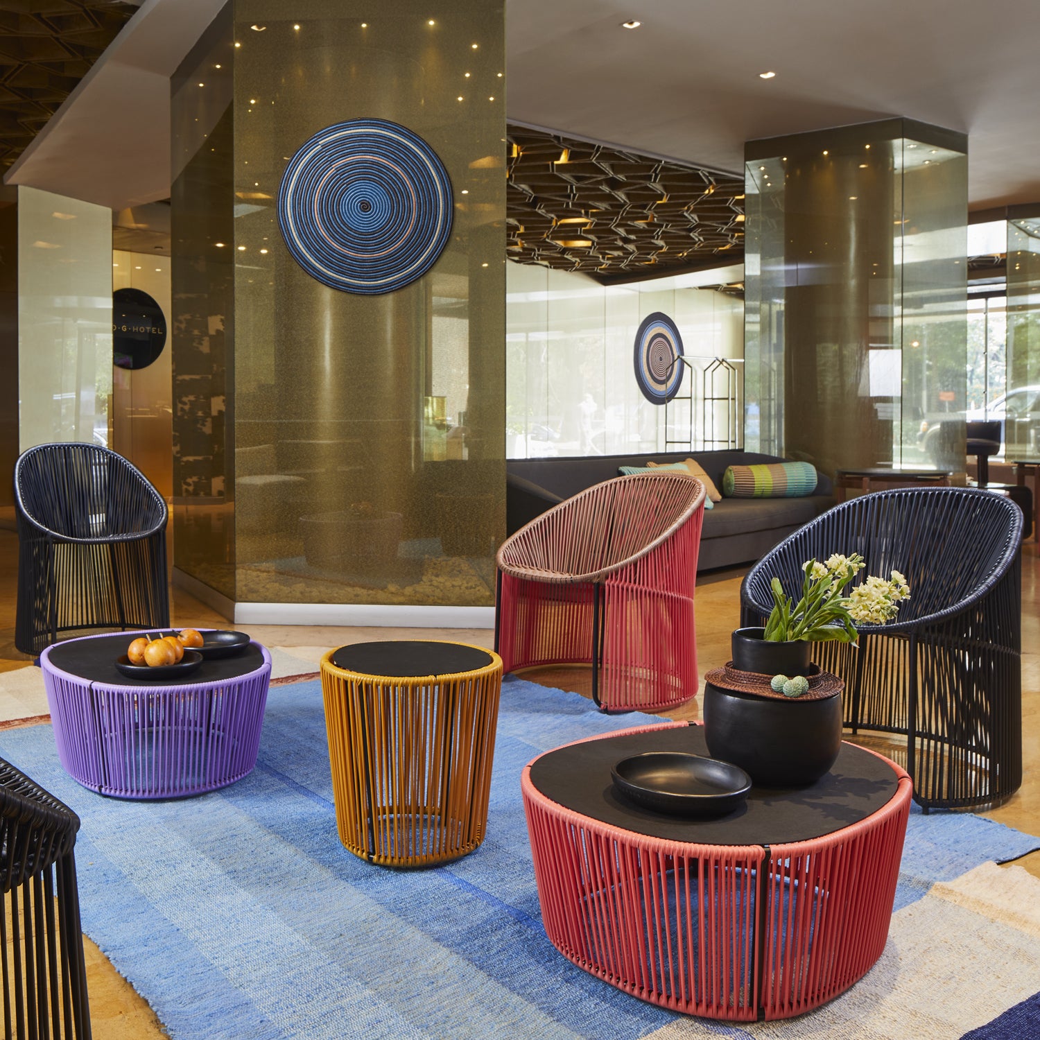 A modern lobby showcases colorful woven chairs and vibrant blue, orange, and red round tables. Ames Cartagenas Sidetable complements the space with decorative items and lush plants. A soft blue rug ties it all together, adding a touch of Sebastian Herkners artistic flair.