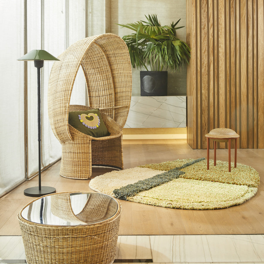 A cozy room featuring Ames’s Cartagenas Natural Coffee Table by Sebastian Herkner—a round, mirror-topped wicker piece—paired with a tall wicker chair, small stool, earth-tone rug, floor lamp, and potted plant against wood and marble walls.