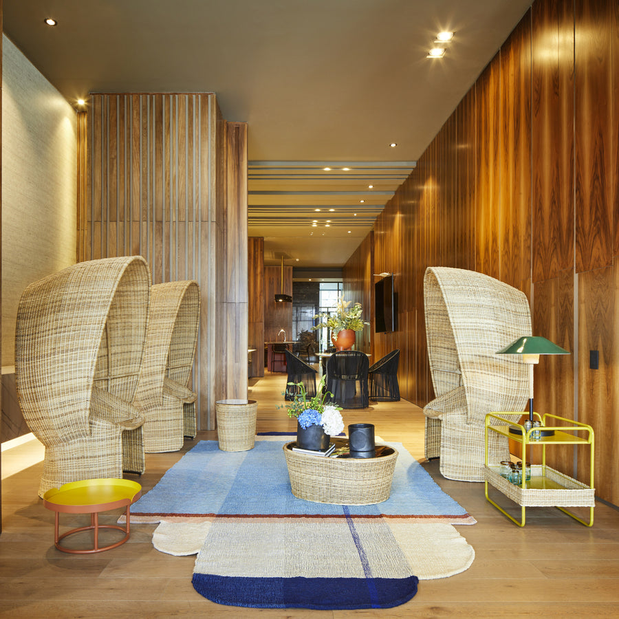 A modern lounge with tall wicker chairs inspired by Ames’ Cartagenas collection, wooden walls, blue patterned rug, yellow side table, decorative plants, and the Ames Cartagenas Natural Coffee Table. Warm lighting adds a cozy, sophisticated touch.