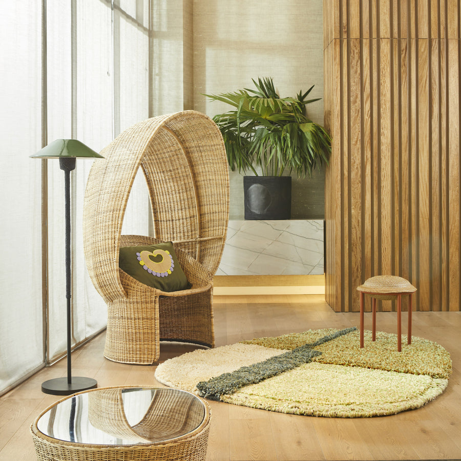 A modern, cozy room features the Ames Cartagenas Natural Reina Chair by Ames—a wicker seat with a green cushion by a black floor lamp. A round rug, small stool, low glass table, and potted plant complete the look against wood walls and large windows.