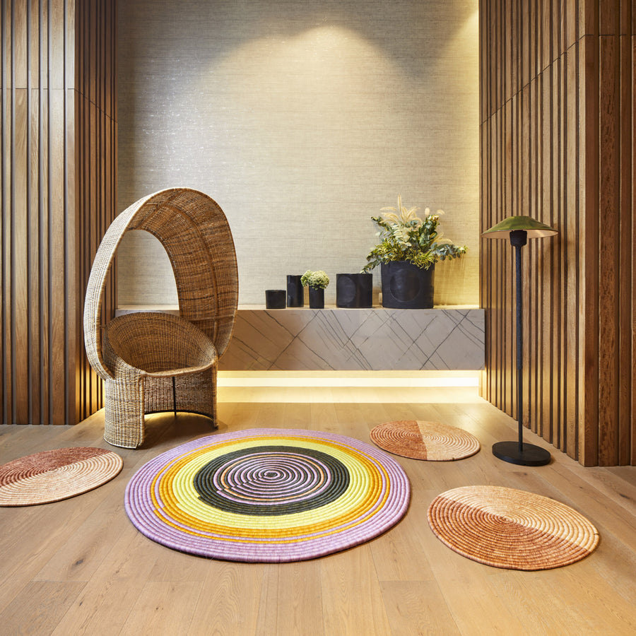 A modern room features an Ames Cartagenas Natural Reina Chair, vibrant circular woven rugs on a wooden floor, a low marble console with black vases and green plants, wood-paneled walls, and soft ambient lighting.