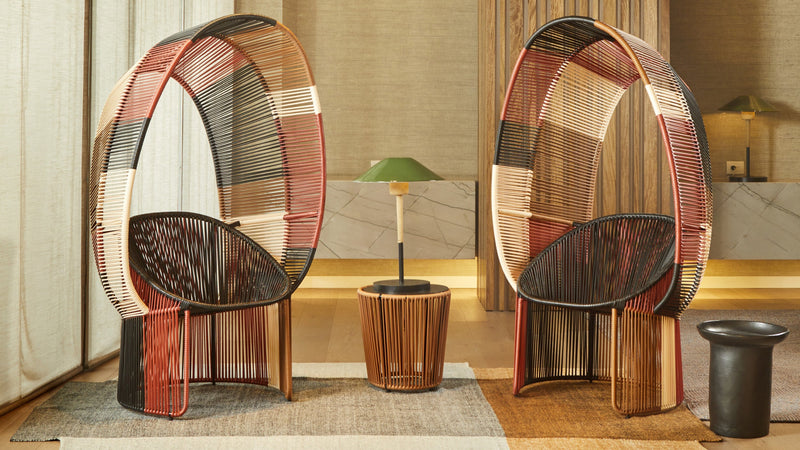 Two Cartagenas Reina Chair Limited Edition wicker chairs from Ames, with multicolored woven backs, enhance a cozy sunlit room. A round side table with a small green lamp sits between them, while a black stool rests on a patterned rug nearby.