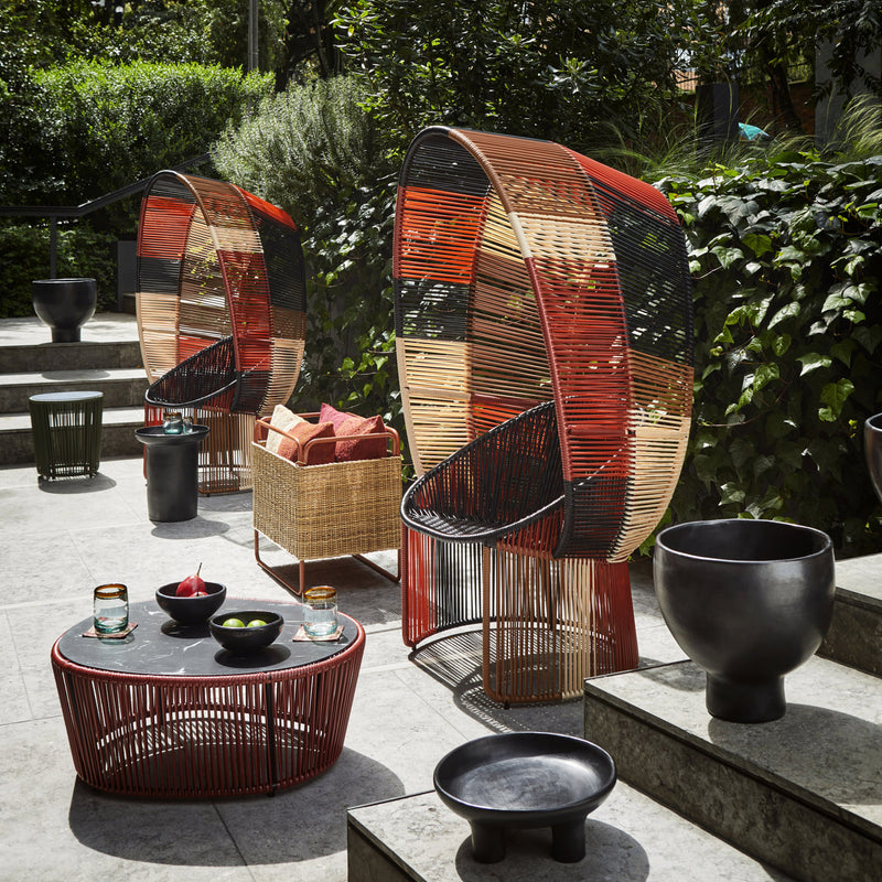 Outdoor patio featuring the Ames Cartagenas Reina Chair Limited Edition with vibrant wicker chairs and round woven tables, complemented by green plants, trees, and decorative bowls and glasses for a charming outdoor setting.