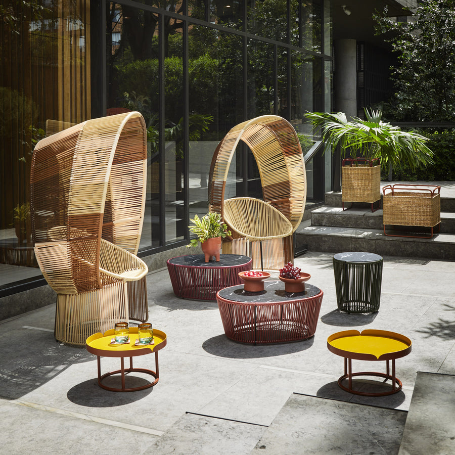The outdoor patio, featuring the Cartagenas Reina Chair Limited Edition by Ames, includes modern wicker chairs and tables with yellow and red accents. Two high-backed Reina Cocoon chairs flank round tables with yellow trays, while a central coffee table displays fruit and small plants amidst lush greenery.