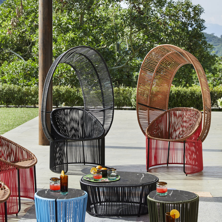 Cartagenas Reina Chairs by Ames, in black, red, and orange with curved backs, are arranged around round black and blue tables topped with drinks. This inviting setup graces a patio with lush greenery behind it.