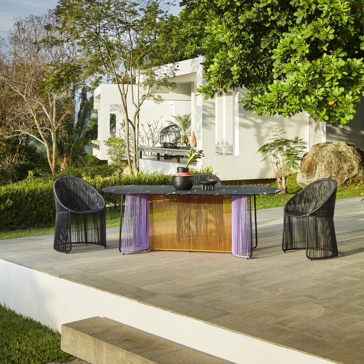 Outdoor patio scene with an Ames Cartagenas Dining Table by Ames, featuring a marble tabletop, two black woven chairs, decorative items, lush green garden, and a modern white building in the background.