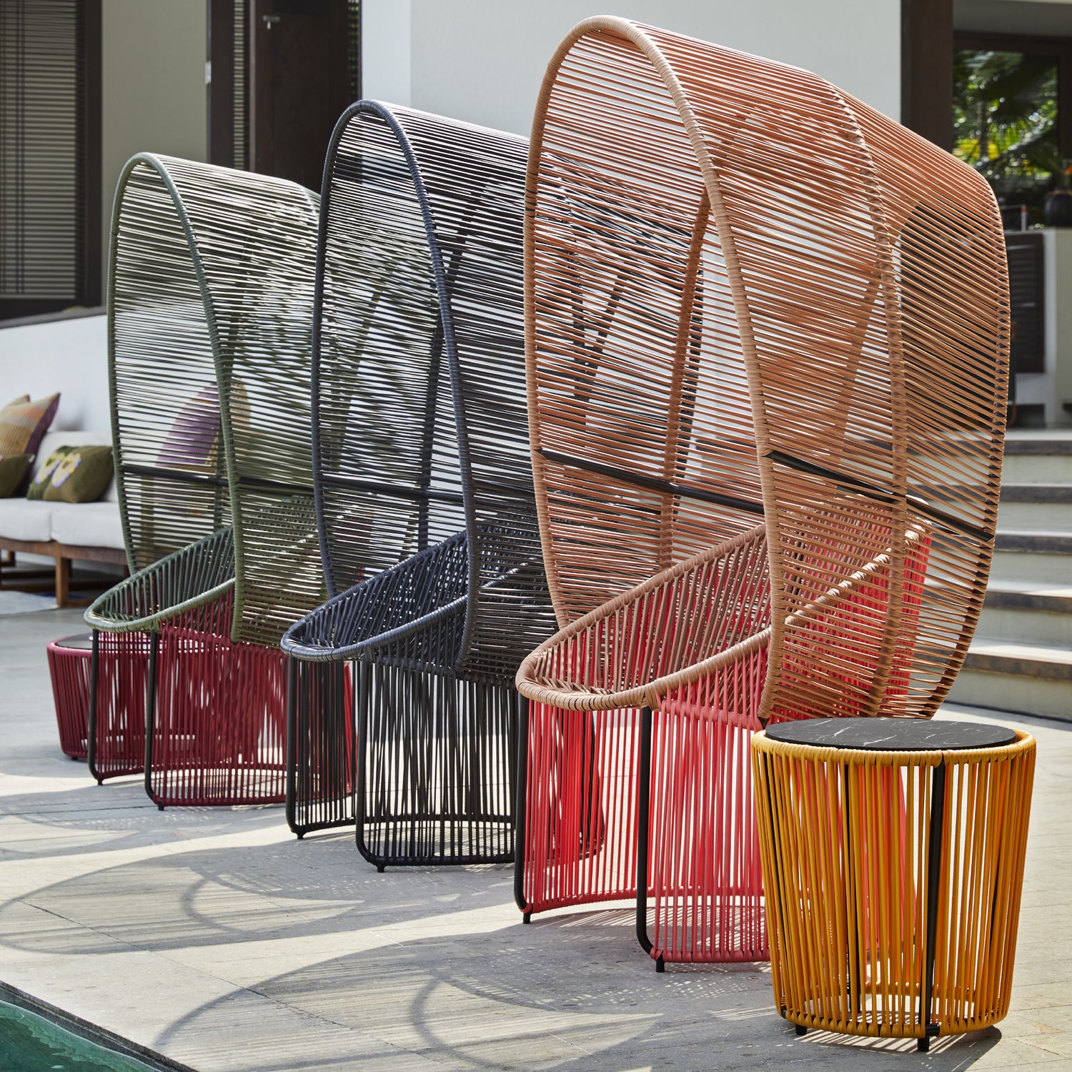 Four Cartagenas Reina Chairs from Ames are arranged poolside, featuring a colorful rounded design made of woven materials in black, orange, and various browns. Coordinated side tables complement the inviting setup, with a staircase and house elegantly gracing the background.