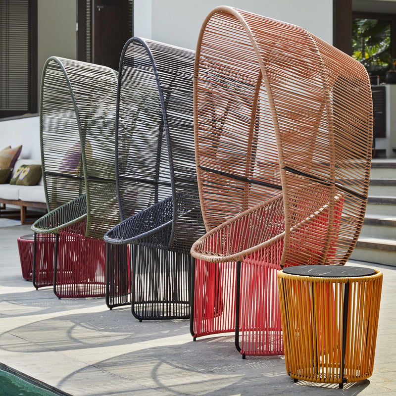 Four vibrant, modern outdoor chairs featuring woven designs in green, black, orange, and red stand by the poolside. Influenced by Sebastian Herkners style, each chair has a tall round back paired with Ames Cartagenas Sidetables. A bustling coastal city with stairs and a building entrance looms behind them.