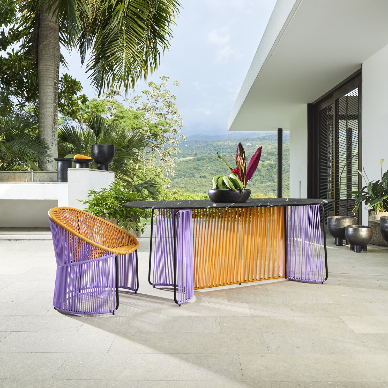 A modern outdoor patio with an Ames Cartagenas Dining Table by Ames, featuring a marble top with tropical fruit, vibrant purple and orange woven chairs, lush greenery, palm trees, and a contemporary white building.