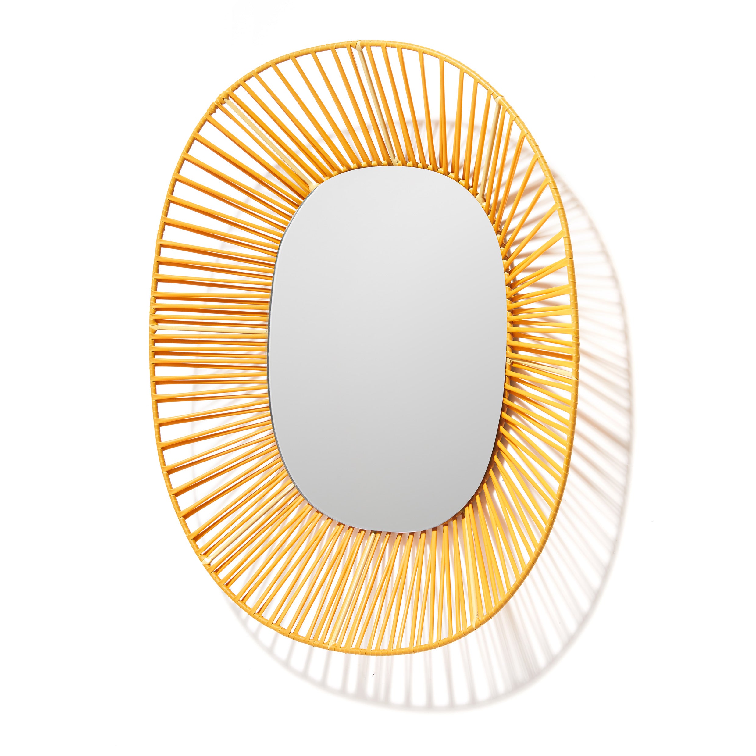 The Cesta Mirror by Ames features an oval shape with a sunburst frame made from yellow rattan and recycled PVC, offering a modern look as shadows are cast on the wall. It stands out against plain white backgrounds, adding vibrant color.