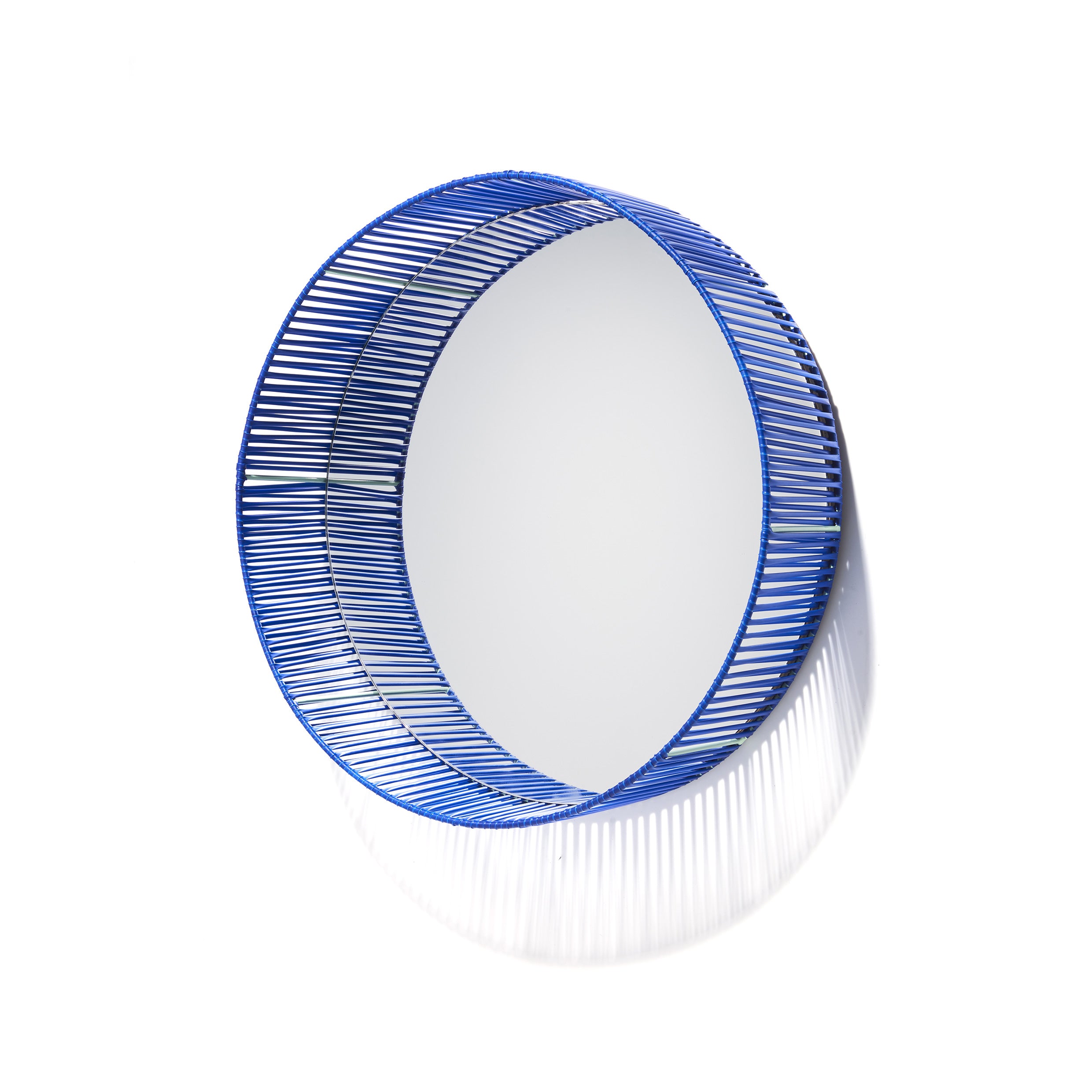 The Cesta Mirror by Ames features a circular design with a blue woven wire frame made from recycled PVC lace, softly shadowed against a white background.