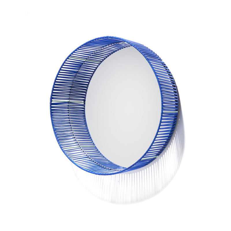 The Cesta Mirror by Ames features a circular design with a blue woven wire frame made from recycled PVC lace, softly shadowed against a white background.