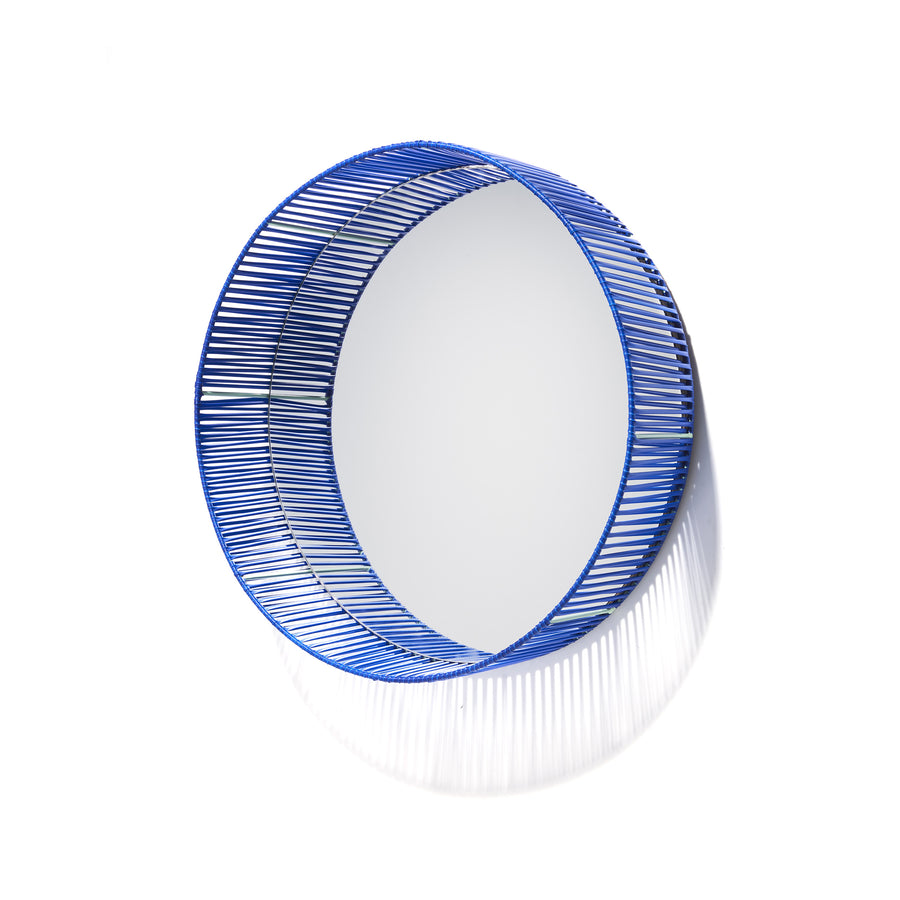 The Cesta Mirror by Ames features a circular design with a blue woven wire frame made from recycled PVC lace, softly shadowed against a white background.