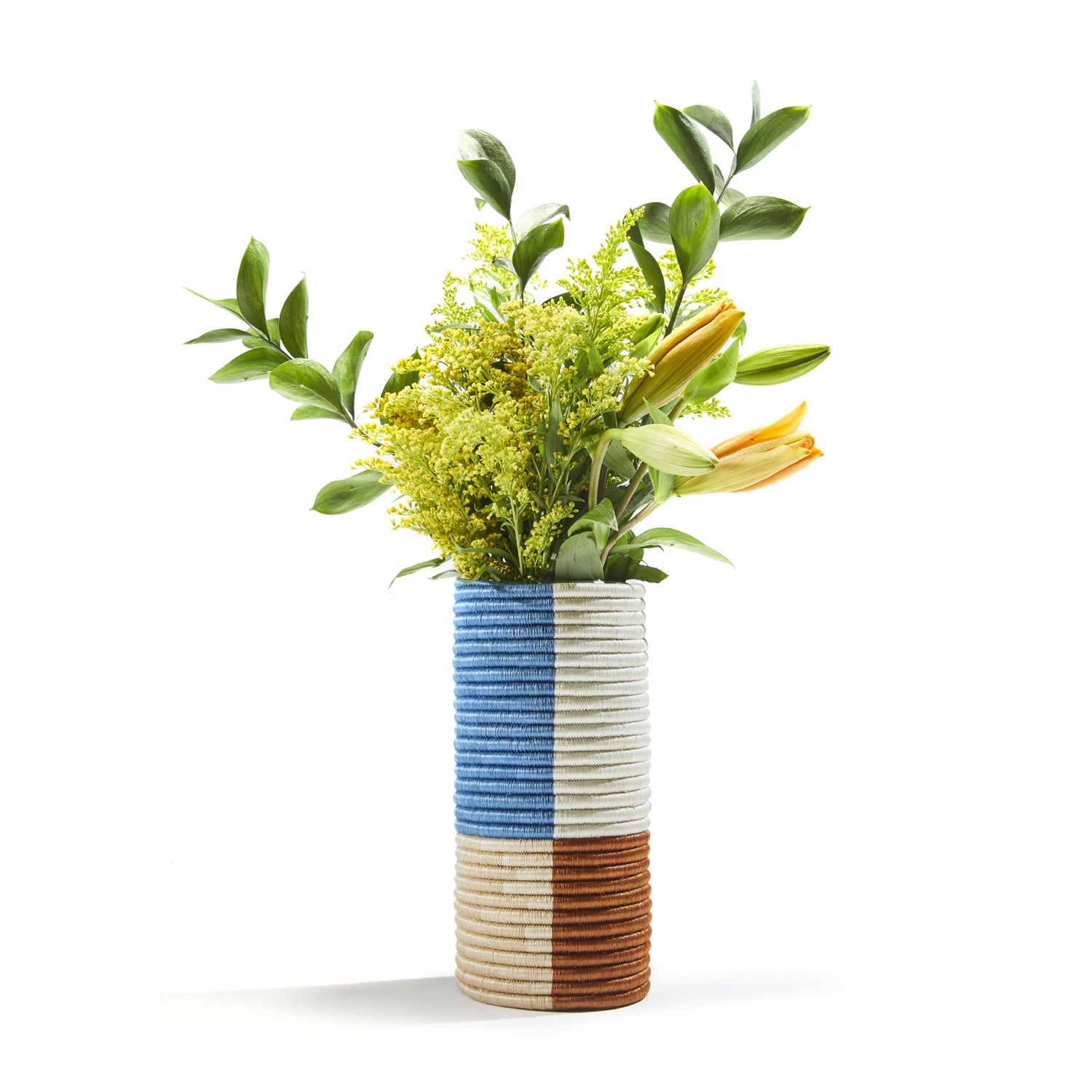 This vibrant cylindrical vase from Ames Choco Vases collection boasts vertical stripes of blue, white, and brown. Expertly crafted with palm fibers for texture, it elegantly displays a bouquet of green leaves, yellow flowers, and an orange lily.