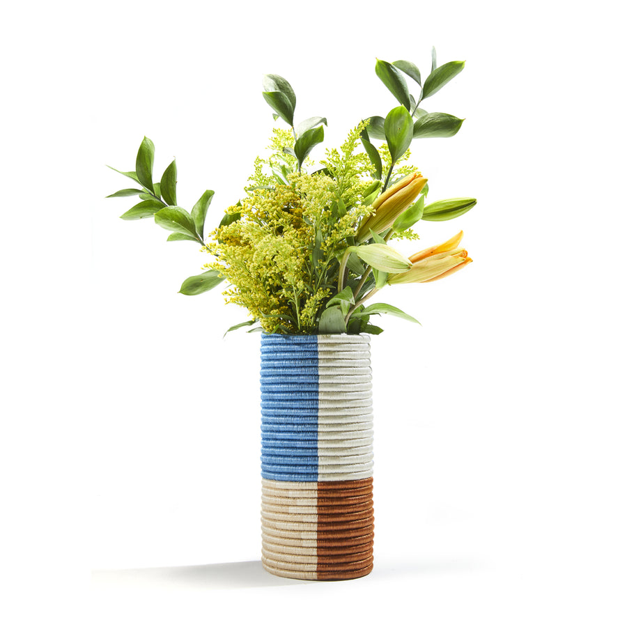 This vibrant cylindrical vase from Ames Choco Vases collection boasts vertical stripes of blue, white, and brown. Expertly crafted with palm fibers for texture, it elegantly displays a bouquet of green leaves, yellow flowers, and an orange lily.