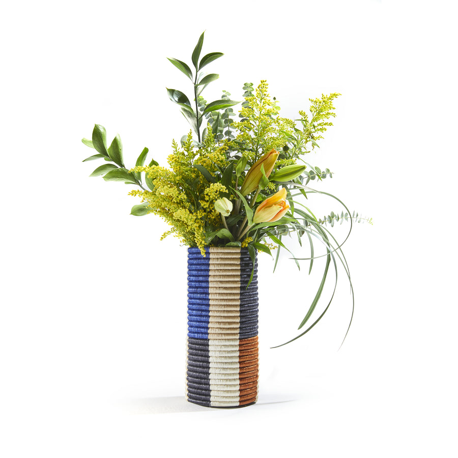 From Ames Choco Vases, this cylindrical vase flaunts blue, white, and brown stripes along with green leaves, yellow flowers, and an orange tulip set against a plain white backdrop.