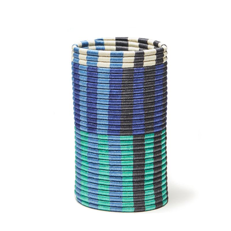 From the Choco Vases collection by Ames, this cylindrical basket is woven with palm fibers and features blue, green, black, and white vertical stripes. Its design includes alternating solid and patterned sections for a vibrant textured look against a white background.