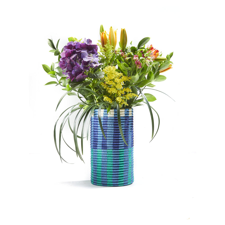 The Choco Vases by Ames display a vibrant bouquet in a blue and green striped vase, featuring elegant purple hydrangeas, orange lilies, yellow flowers, and lush green leaves. The arrangement is crafted with palm fibers for added texture against a white background.