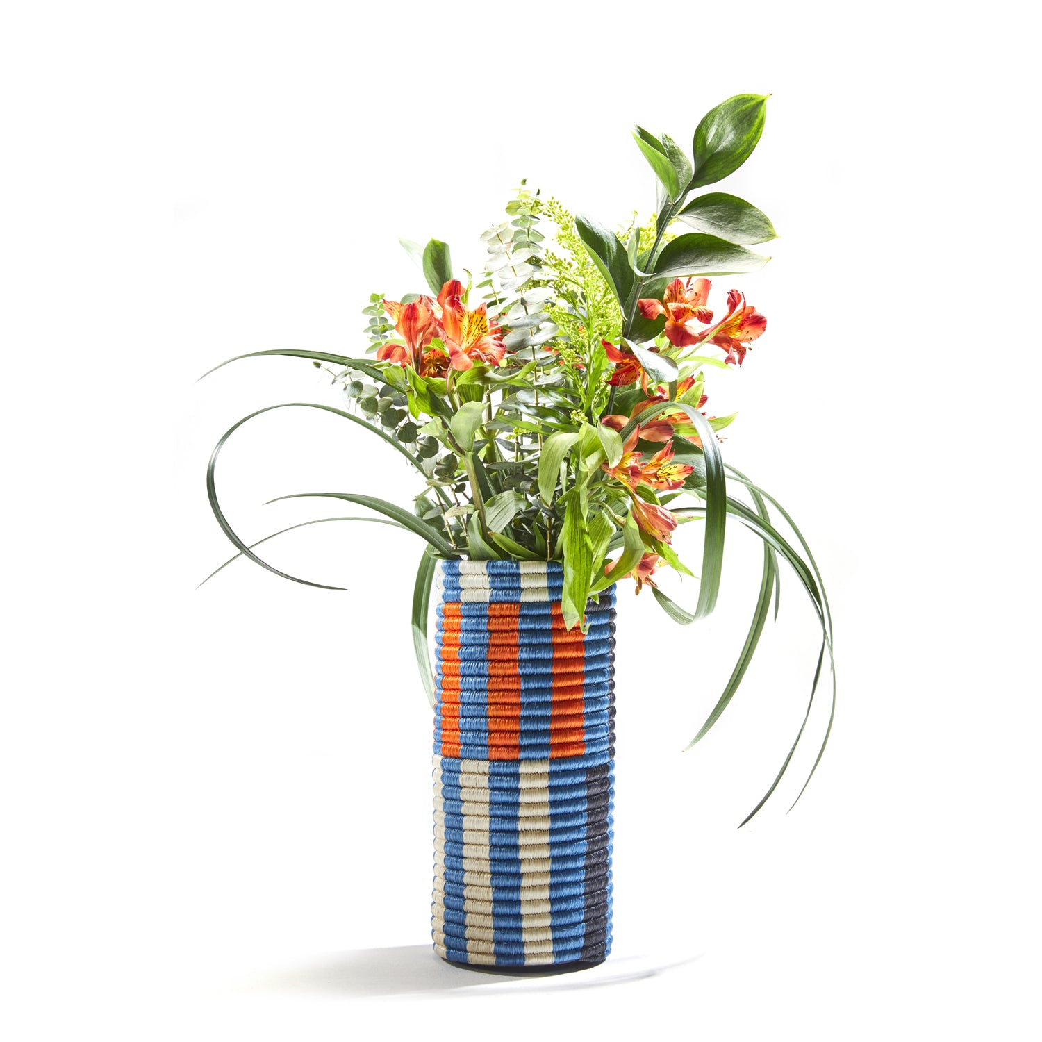 Ames Choco Vases, designed in a tall cylindrical shape with palm fibers, showcase a blue, white, and orange geometric pattern. It elegantly holds vibrant orange flowers, lush greenery, and ferns against a plain white backdrop.