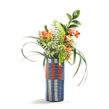 Ames Choco Vases, designed in a tall cylindrical shape with palm fibers, showcase a blue, white, and orange geometric pattern. It elegantly holds vibrant orange flowers, lush greenery, and ferns against a plain white backdrop.