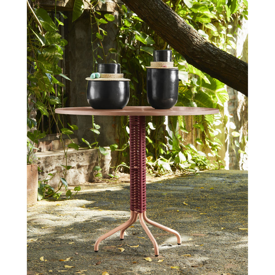 The Cielo Bistro Table by Ames, designed by Sebastian Herkner, features a woven red base and is paired with two black ceramic pots featuring straw accents and lids. The lush green foliage and rustic wall enhance the tranquil scene on this small terrace.