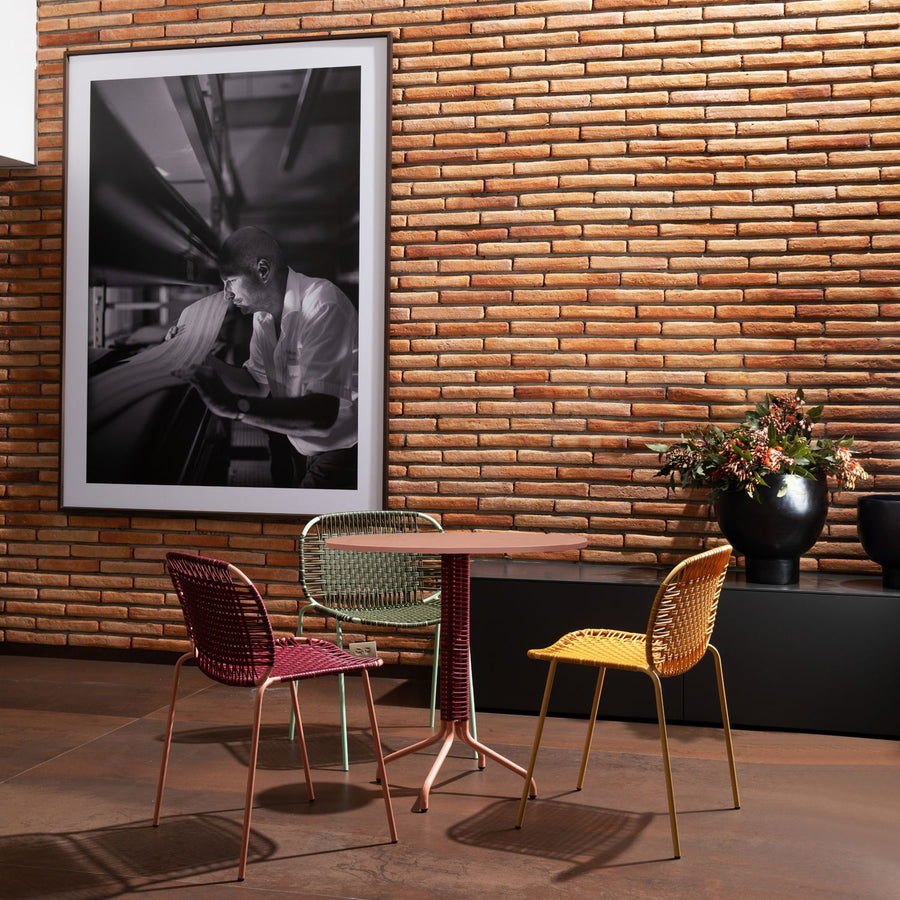 In a modern room with a brick wall, an elegant Cielo Bistro Table by Ames is surrounded by three colorful woven chairs beneath a black-and-white portrait. A shelf with potted plants enhances the compact, harmonious atmosphere.