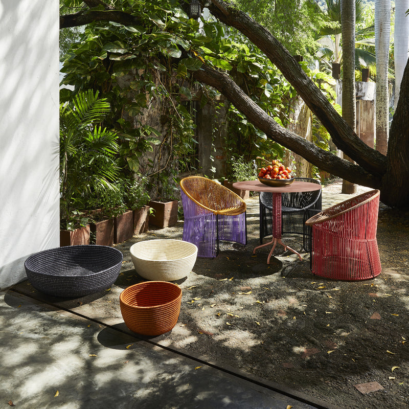 Under a sprawling tree, the cozy outdoor area features Sebastian Herkners compact design with multi-colored woven chairs and baskets around an Ames Cielo Bistro Table holding apples, while lush greenery creates an inviting oasis.