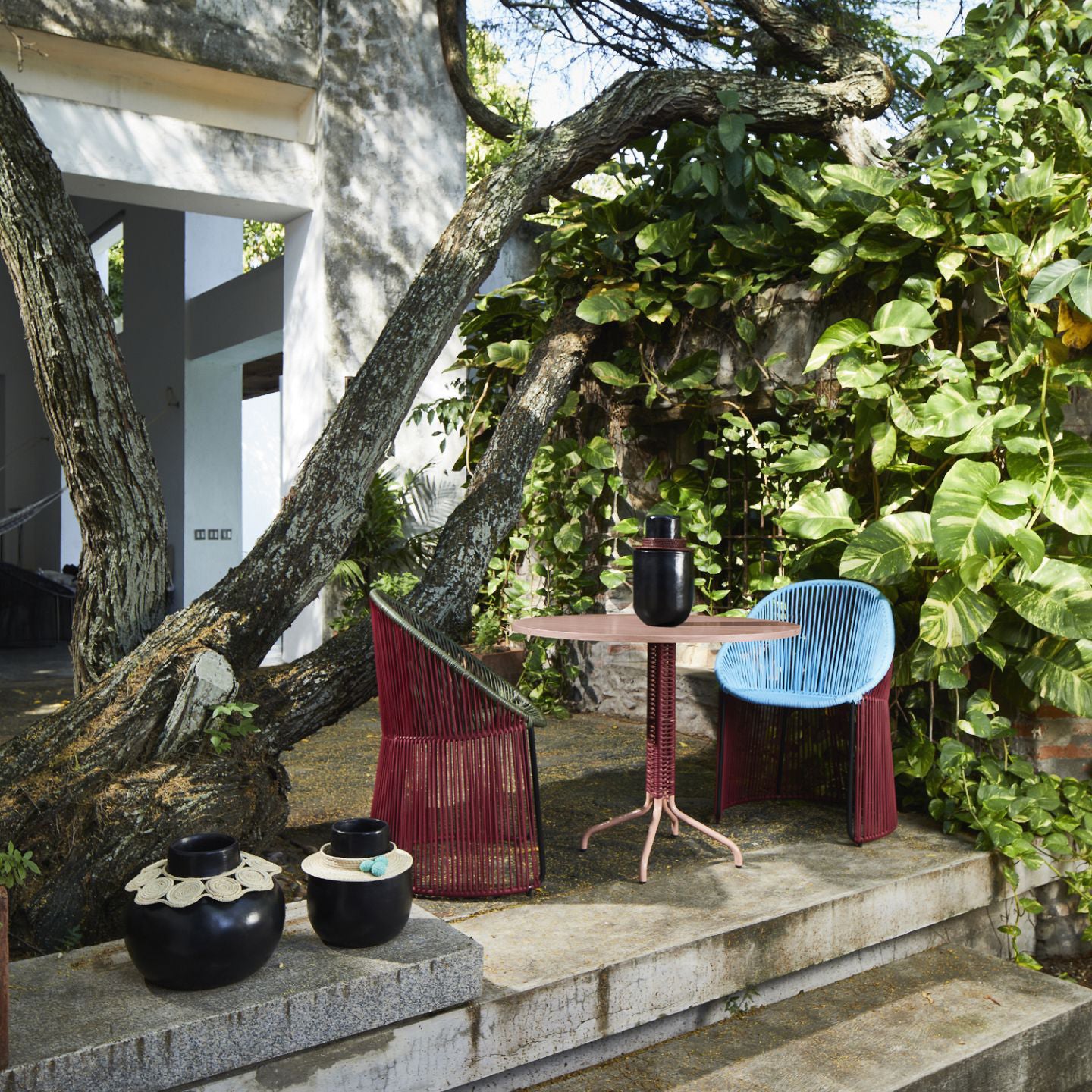 The outdoor scene features a modern patio with colorful chairs by Sebastian Herkner and Ames compact Cielo Bistro Table, decorated with vases. Large tree branches offer shade, while lush green foliage creates a natural backdrop for this stylish furniture arrangement.