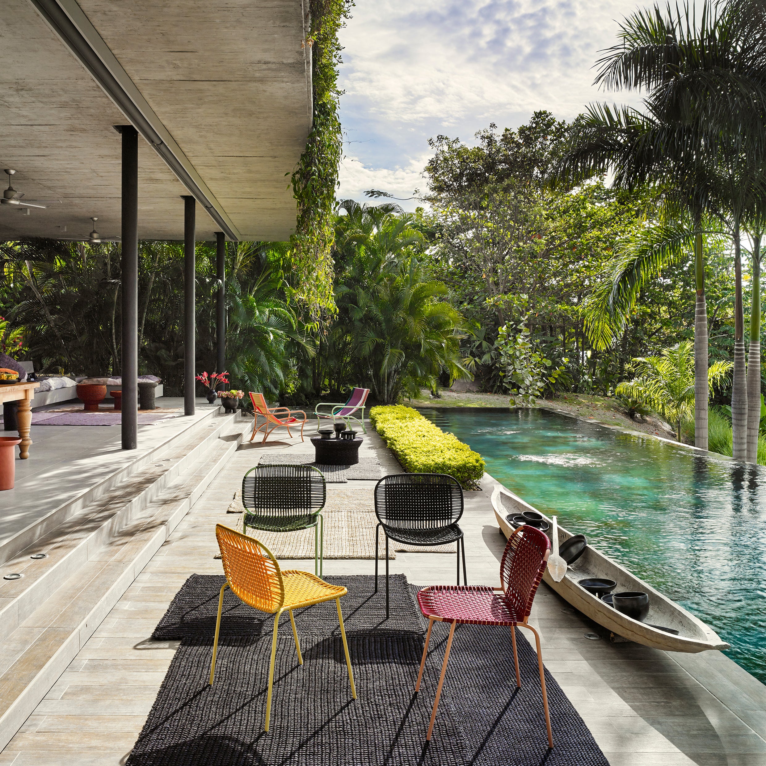 A modern patio with vibrant Ames Cielo Chairs by Ames and bold rugs overlooks a narrow pool bordered by lush tropical plants, where a canoe drifts under tall palms and a partly cloudy sky.