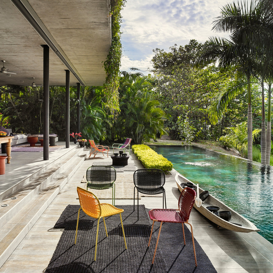 A modern patio with vibrant Ames Cielo Chairs by Ames and bold rugs overlooks a narrow pool bordered by lush tropical plants, where a canoe drifts under tall palms and a partly cloudy sky.