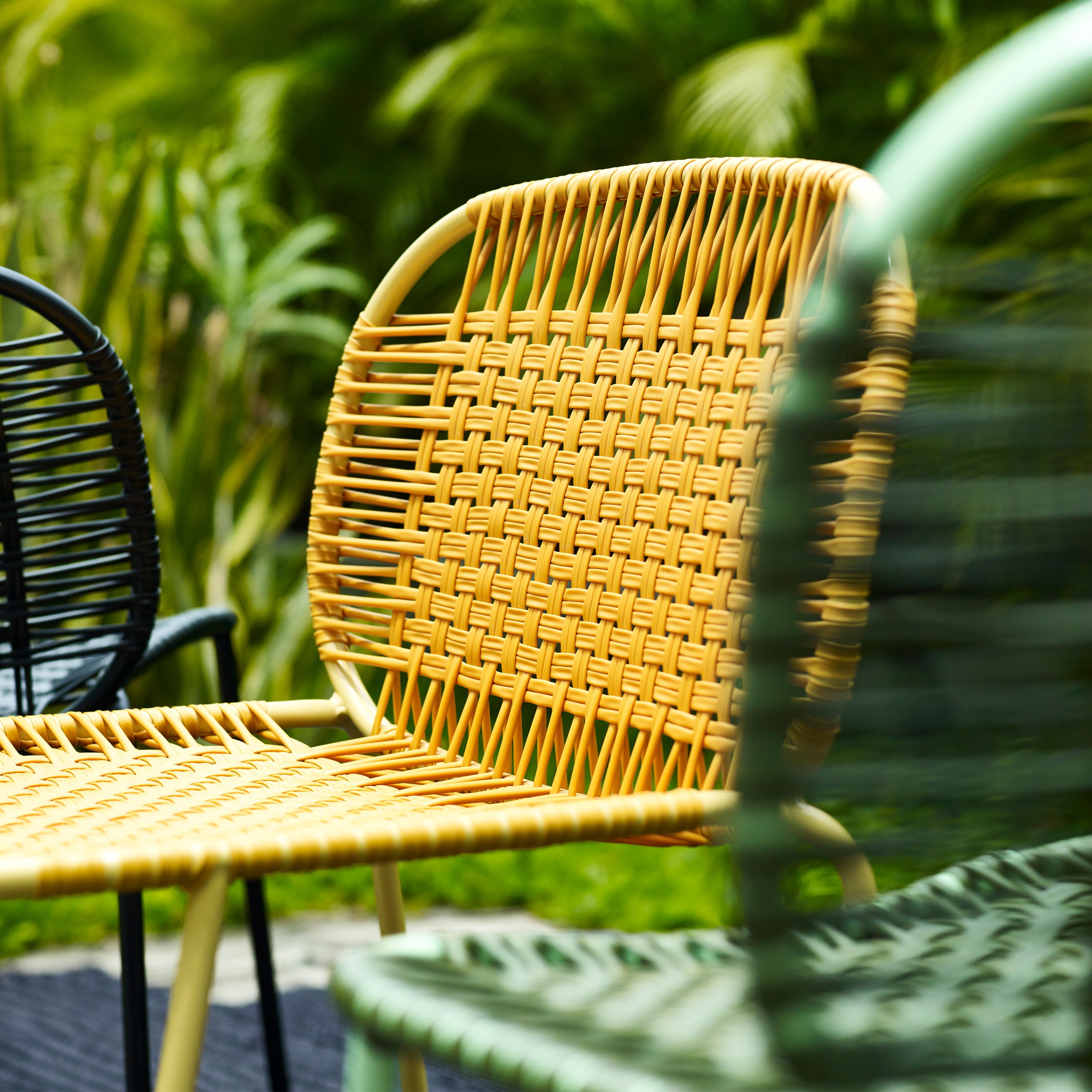 Three Ames Cielo Chairs by Ames, in yellow, green, and black, are arranged outdoors in a lush garden setting with blurred greenery in the background.