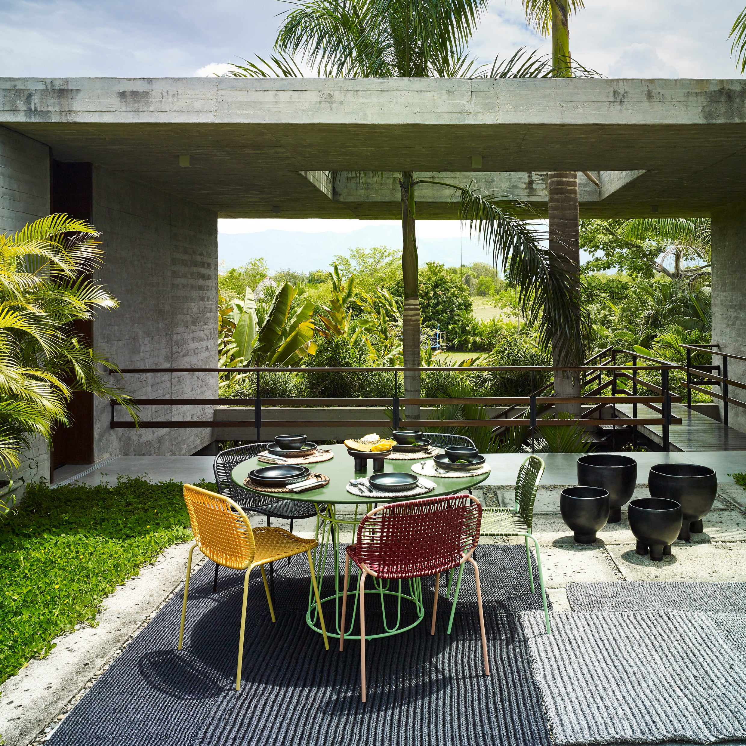 A modern outdoor dining area showcases vibrant Ames Cielo Chairs by Ames around a round table set for a meal, surrounded by lush tropical plants and a concrete structure with large openings.