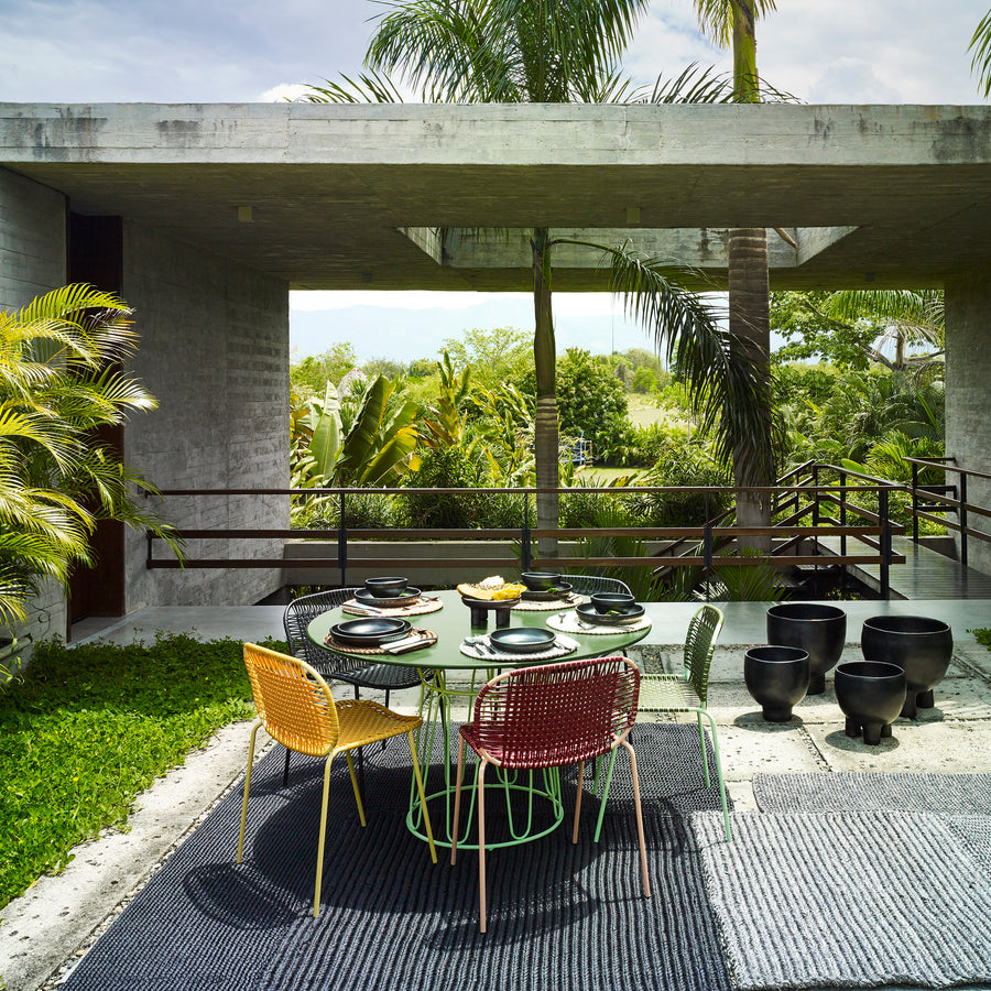 A modern outdoor dining area showcases vibrant Ames Cielo Chairs by Ames around a round table set for a meal, surrounded by lush tropical plants and a concrete structure with large openings.