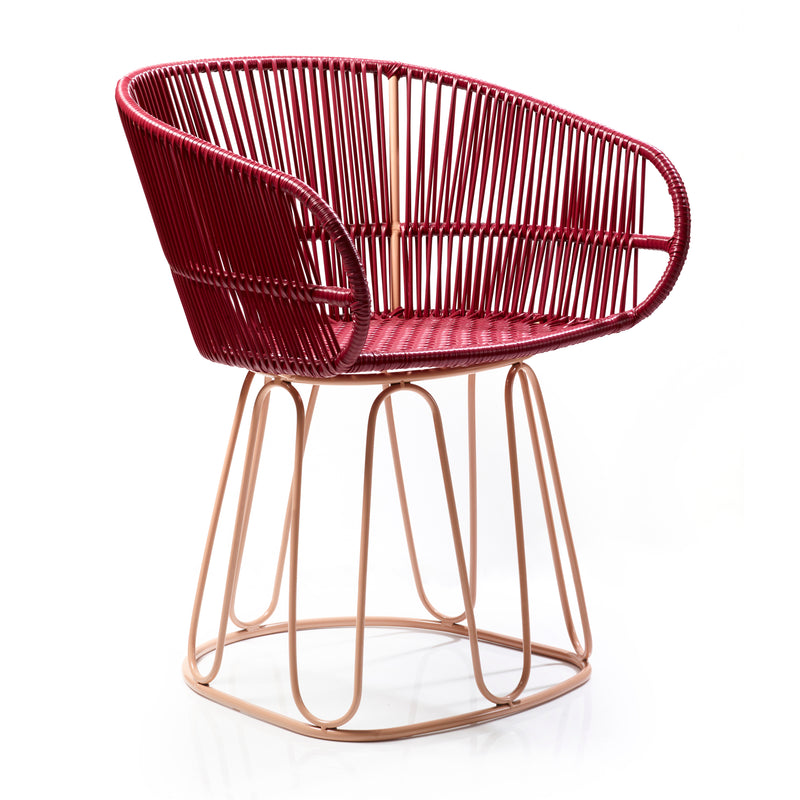This Circo Dining Chair by Ames, handcrafted by Colombian artisans, showcases a red woven design with an open, rounded backrest featuring intricate patterns and a beige metal base with geometric details against a white backdrop.