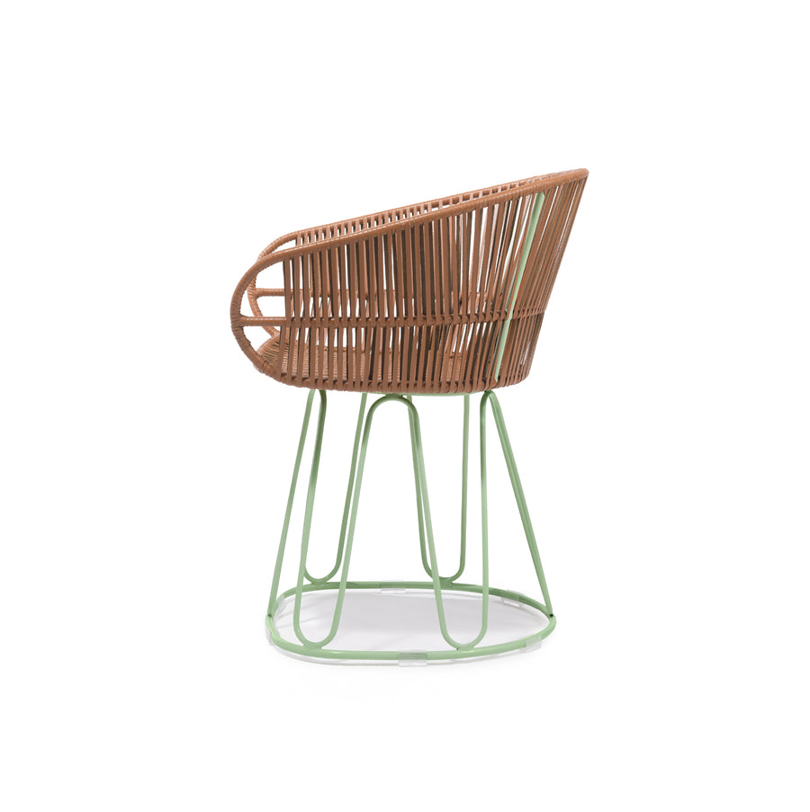 The Ames Circo Leather Dining Chair showcases a contemporary design with a brown woven rattan seat and backrest supported by a green metal frame, featuring curved lines and an open weave pattern that highlights Colombian craftsmanship.