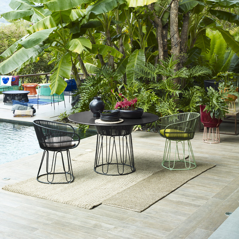 An outdoor patio scene showcases an Ames Circo Leather Dining Table paired with three modern chairs on a woven rug near a pool, surrounded by lush tropical greenery. Decorative accents like a vase and braided leather plant holder adorn the monochromatic tabletop.