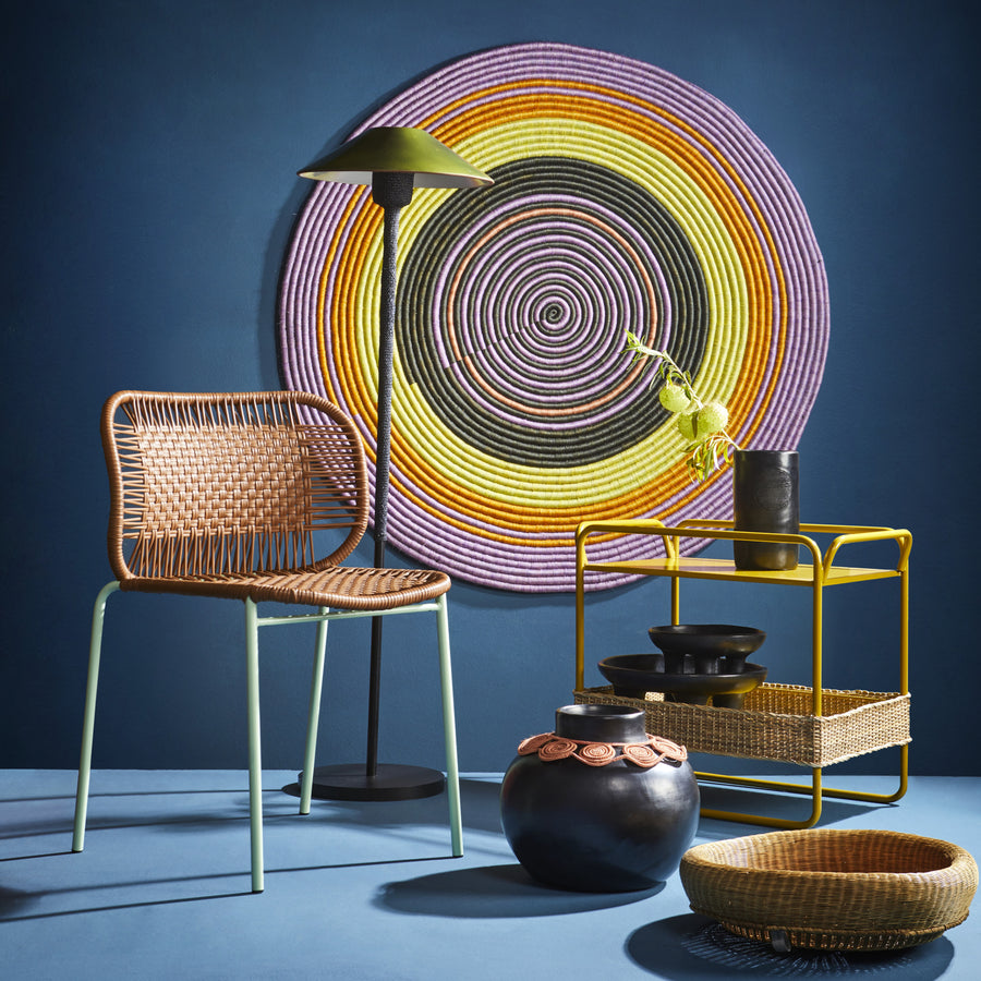 A stylish interior showcases a woven chair, black floor lamp, and vibrant circular wall art. A yellow shelf with a black vase and two bowls sits peacefully. Black pottery is next to a woven basket on the floor, all set against a dark blue backdrop complemented by Ames versatile Dichas Sidetable.