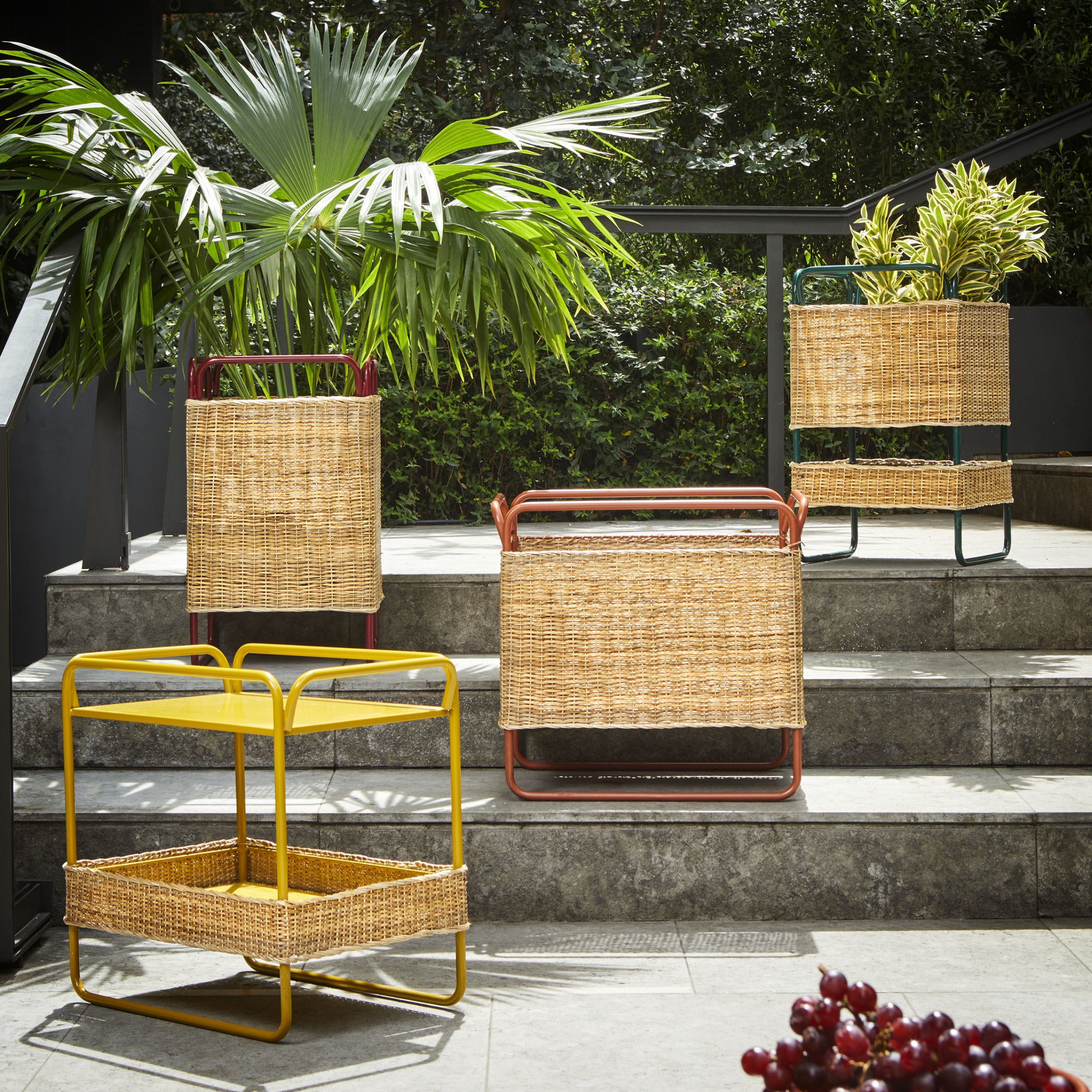 Three wicker plant stands, with frames in recycled plastic—yellow, red, and green—adorn the outdoor steps. A palm and other foliage fill the baskets as sunlight highlights the lush scenery, subtly echoing the vivid tones of Ames Dichas Sidetable.